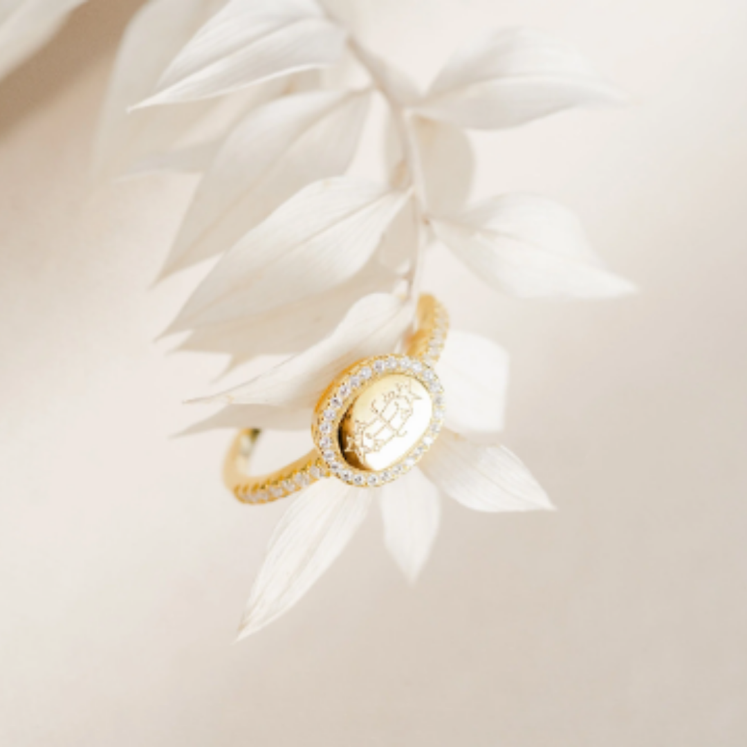gold oval bahai ringstone symbol ring with halo of round diamonds hanging off a white plant with cream background