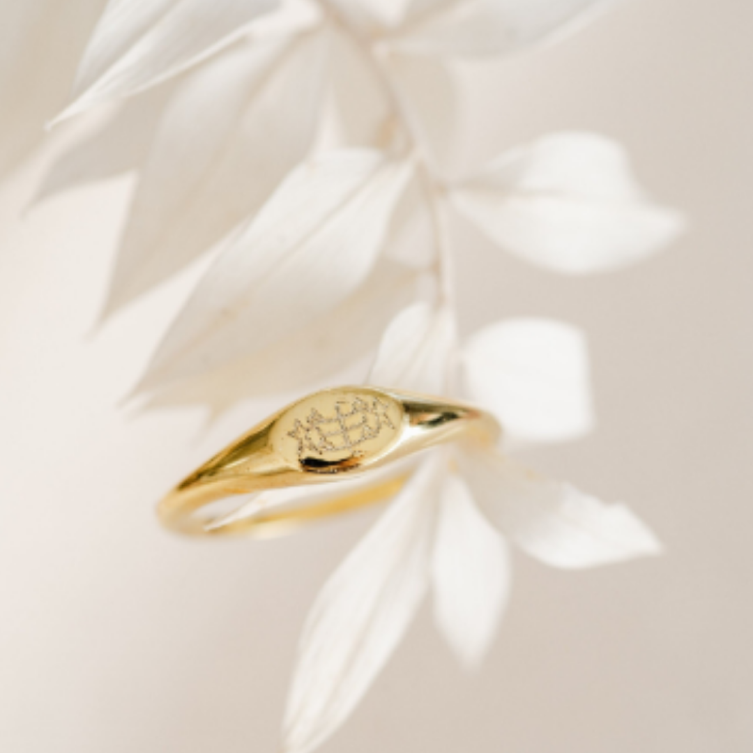 plain and simple gold bahai ring with ringstone symbol engraved on a horizontal oval signet ring draped on a white plant with cream background