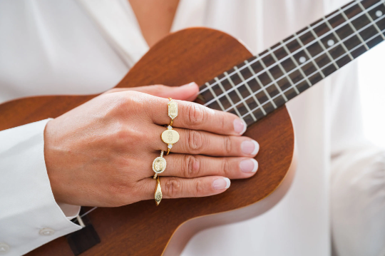 comparison of gold bahai ringstone symbol rings on each finger of a women's hands holding a brown wooden dining table  while holding a brown wooden ukulele 